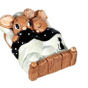 May include: Two brown mice are snuggled together in a bed with a black and white polka dot blanket. The bed has a wooden frame and a black silhouette of a cat on the side.