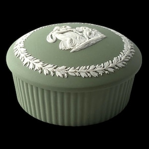 Puede incluir: Una caja redonda de color verde salvia con tapa y adornos en relieve blanco. La tapa muestra una escena clásica central rodeada por una corona de laurel detallada. La base del recipiente presenta un diseño de estrías verticales.