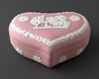 Puede incluir: Un joyero rosa en forma de corazón con adornos en relieve blancos. La tapa muestra una escena clásica con una figura, un caballo y un carro, rodeada por un borde de guirnalda floral y pequeños detalles florales en los lados de la base.