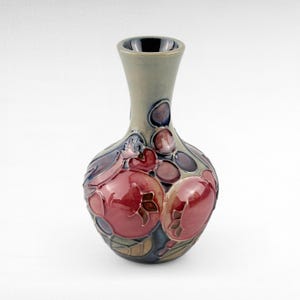 May include: A ceramic vase with a bulbous body and a slender neck. The vase features a design of red pomegranates and blue and purple accents against a muted green background. The vase is approximately 20 cm tall.