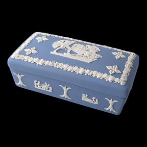 May include: A rectangular, light blue decorative box with white relief detailing. The box features floral and classical relief designs, including a central scene with figures and a horse-drawn chariot. The box is set against a black background.