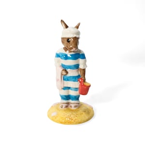 Pode incluir: Uma estatueta de cerâmica de um coelho marrom usando um gorro branco e um pijama listrado azul e branco. O coelho segura um balde vermelho e uma toalha rosa. A estatueta está em uma base amarela e bege.