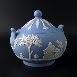 Puede incluir: Un azucarero azul claro con tapa y adornos en relieve blancos. La superficie muestra representaciones blancas en relieve de árboles, follaje y figuras con vestimenta clásica. La tapa tiene un pomo esférico y patrones de hojas.