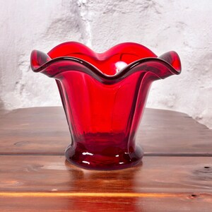 May include: A red glass vase with a ruffled rim. The vase is sitting on a wooden surface.