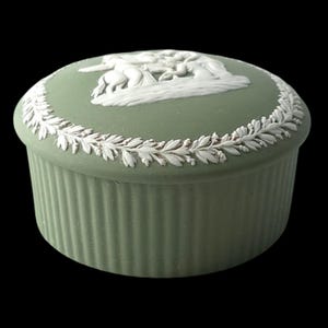 Puede incluir: Una caja redonda con tapa de color verde salvia que presenta un diseño en relieve blanco en la parte superior. La tapa muestra una escena central de figuras y un caballo, rodeada por un borde de hojas de laurel blancas. La base es estriada.