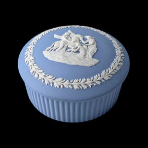 Puede incluir: Caja de baratijas de cerámica azul claro con un diseño en relieve blanco en la tapa. La tapa presenta una escena con figuras y un borde de corona de laurel. La caja tiene una base acanalada y un fondo negro.