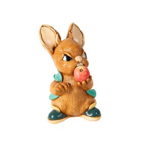 Könnte beinhalten: Eine braune Hasenfigur mit wütendem Gesichtsausdruck, die einen roten Apfel hält. Der Hase trägt eine blaue Weste und grüne Schuhe. Die Figur hat eine detaillierte Fellstruktur und steht vor weißem Hintergrund.