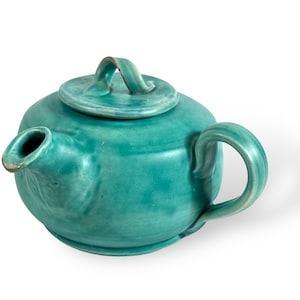 Può includere: Una teiera in ceramica color verde acqua con corpo arrotondato, coperchio con manico e beccuccio. La teiera ha una finitura liscia e smaltata.