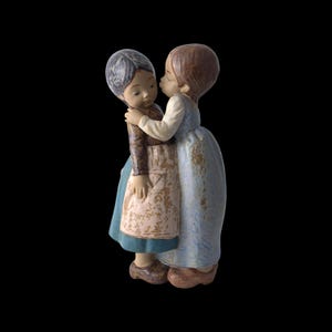 Peut inclure: Figurine en céramique représentant deux jeunes filles s'embrassant. L'une, vêtue d'une robe bleue, embrasse la joue de l'autre. La seconde porte une veste marron, un tablier blanc et une robe bleue. La figurine a une finition mate.