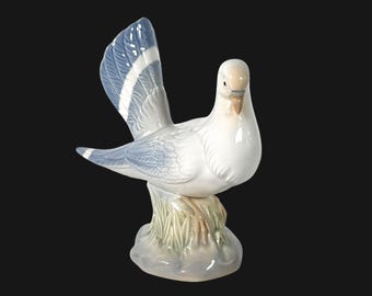 May include: A white porcelain dove figurine with blue and white wings. The dove is perched on a green base with brown accents.
