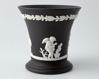 May include: A matte black ceramic vase featuring white relief ornamentation. The upper rim is decorated with a continuous oak leaf and acorn garland. The central body displays a white molded figure of a winged cherub standing by a small urn.