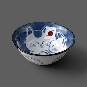 May include: A white ceramic bowl featuring blue hand-painted illustrations of cats. The interior rim has a dark blue textured pattern. A small square sticker is attached to the inner wall with the text MADE IN JAPAN FABRIQUE AU JAPON.