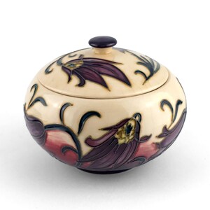 Peut inclure: Une boîte à bijoux ronde en céramique de couleur crème avec un couvercle. La boîte est ornée de motifs floraux stylisés violets et rouges et d'accents vert foncé. Le couvercle a un petit bouton violet foncé.