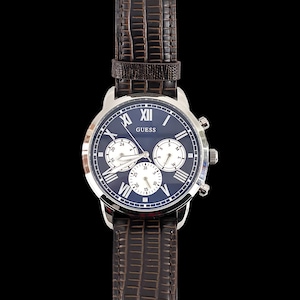 Könnte beinhalten: Eine Guess-Uhr mit dunkelblauem Zifferblatt, silberfarbenem Gehäuse und braunem Lederarmband. Die Uhr hat römische Ziffern, drei Hilfszifferblätter und das Guess-Logo. Das Armband hat ein strukturiertes Krokodilmuster.