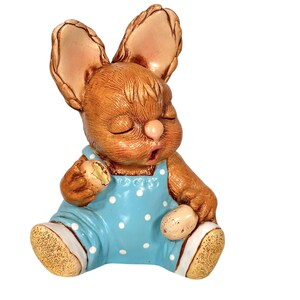 Könnte beinhalten: Eine braune Keramik-Hasenfigur, die eine blaue Latzhose mit weißen Punkten trägt. Der Hase hält ein kleines braunes Ei in der einen Hand und ein größeres braunes Ei in der anderen.