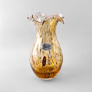 Puede incluir: Un jarrón de vidrio transparente y color ámbar con borde ondulado y patrón moteado. Una pegatina circular en el frente dice: VETRO ESEGUITO SECONDO LA TECNICA DEI MAESTRI DI MURANO MADE IN ITALY. Se incluye una pequeña pieza de vidrio millefiori incrustada.