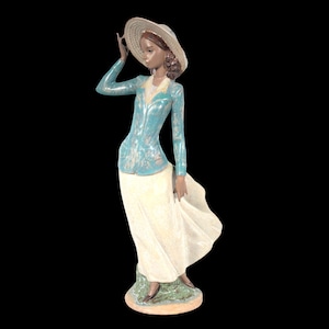 May include: A porcelain figurine of a woman wearing a blue and white patterned dress and a straw hat. She is standing on a base with green and brown accents.