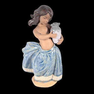 May include: A ceramic figurine of a young person holding a small vase. The figure has brown hair and is wearing a blue skirt with white trim. The vase is white with a gray top. The figurine is on a beige base.