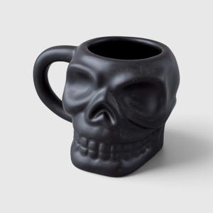 May include: A matte black ceramic mug shaped like a human skull. The mug features a hollow interior, a curved handle on the left side, and moulded facial details including deep eye sockets, a nasal cavity, and a row of teeth along the lower edge.