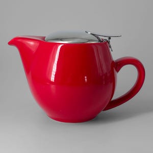 Op de afbeelding: Een felrode keramische theepot met een rond lichaam, een gebogen handvat en een puntige tuit. De theepot is voorzien van een gepolijst zilverkleurig metalen deksel met een scharnier en een platte hendel aan de bovenkant om hem te openen.