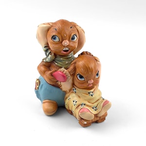 May include: A ceramic figurine depicting two brown bunnies. One bunny is standing and holding a pink brush, wearing blue overalls and a green scarf. The other bunny is seated, wearing a yellow dress with blue floral accents.