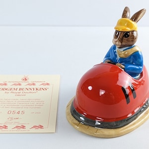 May include: A red and white dodgem car with a brown bunny wearing a blue shirt and yellow hat driving it. The figurine is part of the 'Dodgem Bunnykins' collection by Royal Doulton. The text on the base reads 'DB249'.