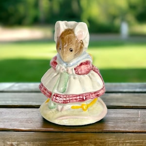 May include: A ceramic figurine of a mouse wearing a white dress with a red and white plaid trim, a green and white scarf, and a white bonnet. The mouse is holding a green and white object in its paws.