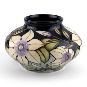 Op de afbeelding: Een keramische vaas met een donkerblauwe achtergrond en een bloemenontwerp. De bloemen zijn wit met paarse centra, en de bladeren zijn groen en bruin. De vaas heeft een afgeronde vorm en een zwarte rand.