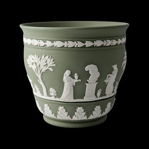 May include: A sage green ceramic planter with white bas-relief designs. The planter features a repeating leaf pattern at the top and bottom, with a central band depicting classical figures and a tree. The planter has a rounded shape.