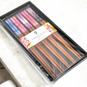 May include: A set of five pairs of chopsticks with floral patterned tops in a black box. The chopsticks are made of wood and have a brown finish. The box has a label that says "Chopsticks Set" and "Made in Japan".