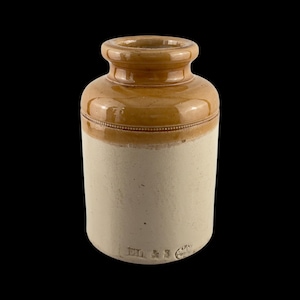 May include: A vintage stoneware jar with a two-tone design. The top section is glazed in a warm brown, while the lower portion is a creamy off-white. A decorative band encircles the jar. The base has stamped text and a circular emblem.