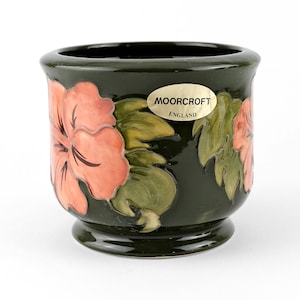 May include: A dark green ceramic pot with a floral design. The pot features large, stylized pink flowers and green leaves. The pot has a small base and a rounded shape. The text "MOORCROFT ENGLAND" is on a sticker on the pot.