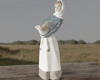May include: A ceramic figurine depicting a woman cradling a lamb. The woman is dressed in a white headscarf, a grey-blue top, and a long white skirt with a grey hem. The lamb is light brown. The background is a blurred outdoor scene.