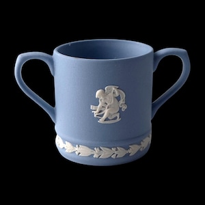 Peut inclure: Une tasse en céramique bleue avec deux anses, ornée d'un motif en bas-relief blanc représentant une figure et d'une bordure de feuilles décoratives. La tasse est posée sur un fond noir.