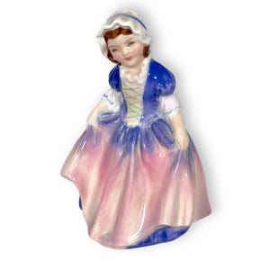May include: A porcelain figurine of a girl wearing a blue and pink dress with a white bonnet. The figurine is in a vintage style.