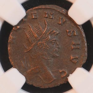Puede incluir: Una moneda romana de bronce, con el perfil de un hombre con una corona de laurel e inscripciones. La moneda está en un soporte transparente con clips blancos, sobre un fondo negro.