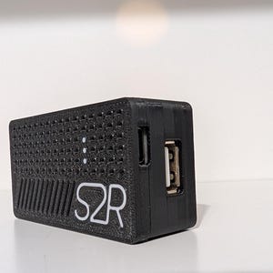 Może przedstawiać: Czarne prostokątne urządzenie o teksturowanej powierzchni, z portem USB i białym napisem "S2R". Na urządzeniu widoczne są trzy małe białe światełka. Urządzenie jest prawdopodobnie akcesorium elektronicznym.