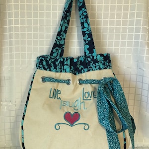 May include: A beige canvas tote bag with blue and teal floral print fabric straps and trim. The bag has a drawstring closure and a blue and teal ribbon. The bag has the text "Live. Laugh. Love." embroidered on the front with a red heart.