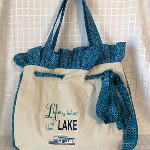 May include: A beige canvas tote bag with blue and white patterned fabric trim. The bag has a drawstring closure and a blue ribbon. The bag has the text "Life is better at the LAKE" embroidered on the front with a small boat image.