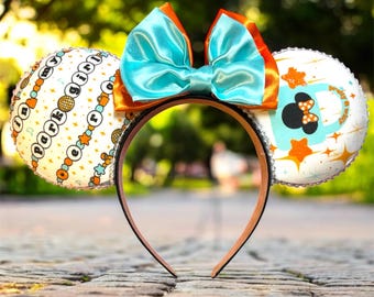 May include: A pair of mouse ears with a teal satin bow and orange accents. The ears are white with a pattern of stars, circles, and the text "I'm my park girl". The headband is brown.