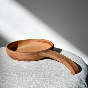 May include: A handmade, terracotta-colored ceramic pan with a long handle. The pan has a shallow, round bowl and a smooth, unglazed surface. The handle is integrated into the design, creating a seamless look. The pan is likely used for cooking or serving.