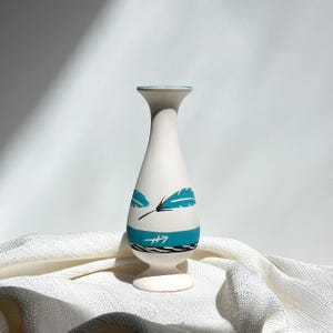 May include: A white ceramic vase with a flared top and a round base. The vase features teal feather designs and a teal band with a stylized wave pattern. The vase is approximately 8 inches tall.