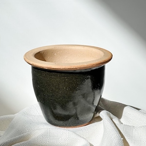 May include: A dark green ceramic planter with a light brown rim. The planter has a rounded shape and a wide opening. The interior is a light beige color. The planter is sitting on a white cloth.