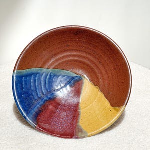 May include: A round ceramic bowl featuring a speckled reddish-brown base glaze. The interior surface is decorated with three distinct, overlapping glazed sections in vibrant blue, deep red, and golden yellow, creating a segmented color pattern.
