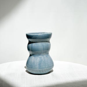 May include: A small, hand-thrown ceramic vase with a glossy, dusty blue glaze. The vessel features a distinct, organic hourglass shape with a wide, rounded base that tapers inward toward the middle before widening slightly at the top rim.