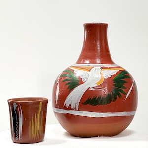 May include: A terracotta-colored clay vase with a rounded body and narrow neck, featuring a hand-painted white bird with green wings. Beside it sits a matching small cylindrical clay cup decorated with vertical white and yellow brushstrokes.