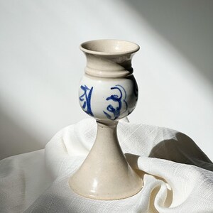 May include: A ceramic vase with a cream-colored base and rim, and a white body adorned with blue abstract designs. The vase has a bulbous midsection and a flared base, standing on a white cloth.