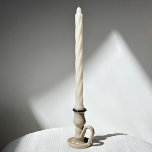 May include: A tall, off-white twisted candle stands in a beige ceramic candlestick with a dark blue accent. The candlestick has a handle and a round base. The candle and holder are on a white surface.