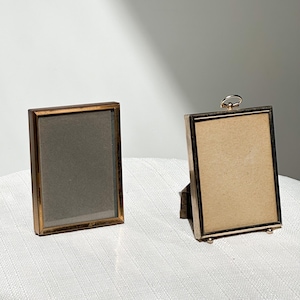 May include: Two rectangular picture frames with gold-colored metal borders. One frame is standing upright with a small stand, and the other is lying flat. Both frames have a glass front and a neutral-colored backing.