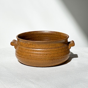 May include: A handmade, brown ceramic bowl with two handles. The bowl has a wide, shallow shape with a textured surface and a dark brown rim. The bowl is likely used for serving food.